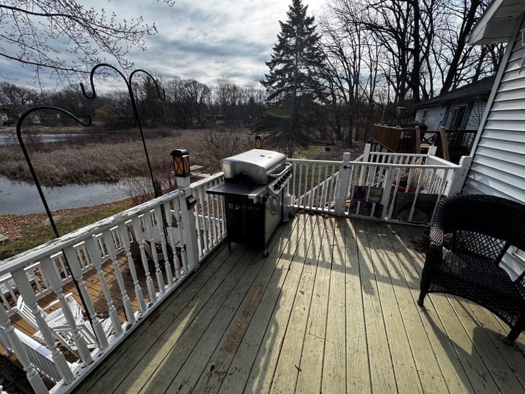 Elevated wood deck with white railing, grill, and patio furniture overlooking a pond